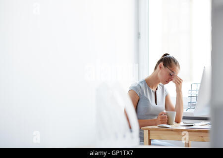 Stanco giovane donna seduta alla scrivania per computer con tazza di caffè toccando la fronte con la sua mano, sentendosi stressati e oberati di lavoro, Foto Stock