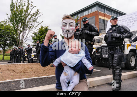 Un uomo che indossa un Guy Fawkes 'Anonimo' maschera, e tenendo un bambino, dà un saluto indifferente di fronte a una fila di PSNI gli ufficiali di polizia Foto Stock