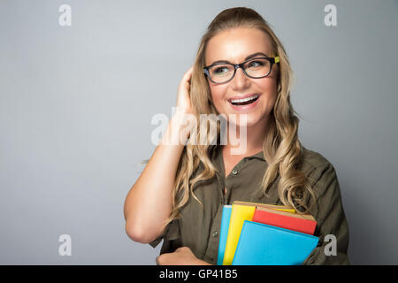 Studente di Cute girl indossando le graminacee in piedi con i libri di testo su sfondo grigio, pronto per la nuova stagione di studio, godendo di istruzione Foto Stock