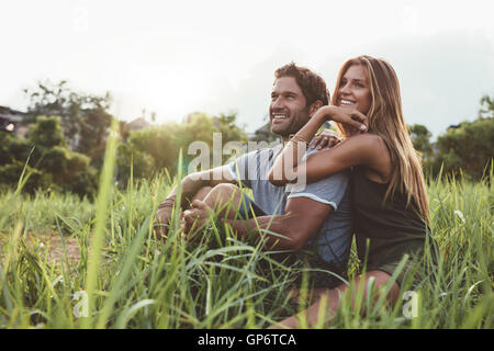 Affettuosa coppia giovane seduto su erba rurale e guardando lontano sorridente. Giovane uomo e donna seduta nel campo guardando a vista. Foto Stock