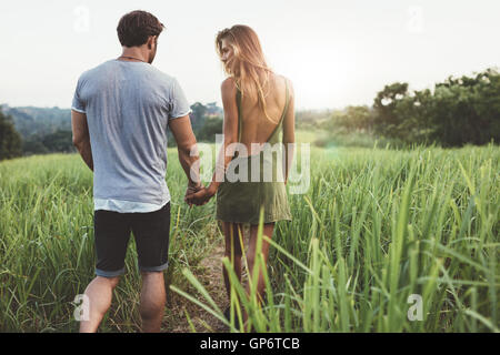 Vista posteriore del tiro del giovane uomo e donna holding hands camminare insieme attraverso il campo di erba. Coppia giovane a piedi attraverso grassy ro Foto Stock