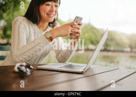 Colpo di donna sorridente tramite telefono cellulare al cafe. Giovane donna seduta a tavola con il computer portatile la lettura di un messaggio di testo sul suo Foto Stock