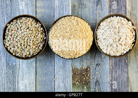 Bocce di lenticchie, bulgur e i fiocchi d'avena in una fila è vista dal di sopra su un vecchio tavolo di legno. Foto Stock