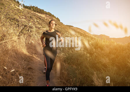 Giovane donna correndo giù per una montagna in piena luce solare con essiccazione erba dietro lei mentre vestita di nero e grigio Foto Stock