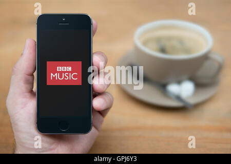 Un uomo guarda al suo iPhone che visualizza la BBC Music logo, mentre sat con una tazza di caffè (solo uso editoriale). Foto Stock