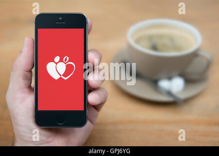 Un uomo guarda al suo iPhone che visualizza il dare la donazione di sangue logo, mentre sat con una tazza di caffè (solo uso editoriale). Foto Stock