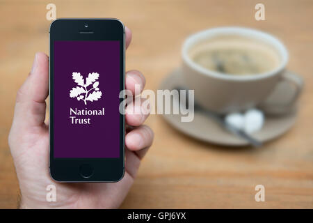 Un uomo guarda al suo iPhone che visualizza il National Trust logo, mentre sat con una tazza di caffè (solo uso editoriale). Foto Stock