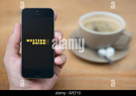 Un uomo guarda al suo iPhone che visualizza la Western Union il logo, mentre sat con una tazza di caffè (solo uso editoriale). Foto Stock