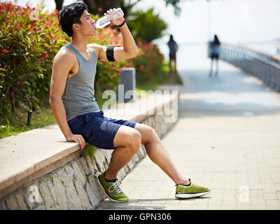 Giovane bello pareggiatore asiatici prendendo una pausa e acqua potabile da una bottiglia, vista laterale Foto Stock