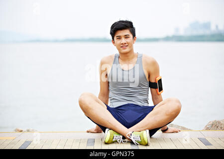 Giovani asiatici bello indossare pareggiatore tracker fitness seduta dal lago guardando sorridente della fotocamera Foto Stock