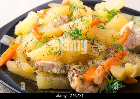 Fette di patate in umido, il pollo, la carota e la cipolla cosparsi di spezie e il verde di aneto su una piastra nera closeup Foto Stock
