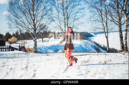 Sorridente giovane bella donna bionda in inverno russo suit corre attraverso i cumuli di neve. Ragazza vestita sciarpa, rivestire di pelle di pecora e luminose lungo mantello rosso Foto Stock