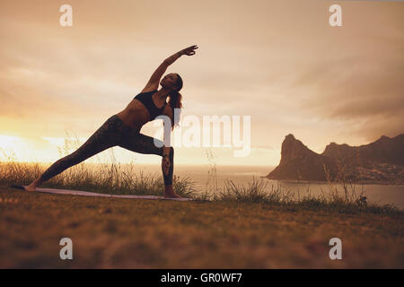 A piena lunghezza shot della giovane donna pratica Utthita Parsvakonasana Yoga asana. Femmina sano esercizio al tramonto sulla scogliera. Foto Stock