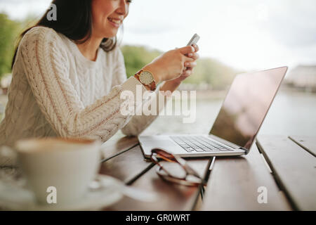 Ritagliato colpo di giovane donna utilizzando il telefono cellulare a outdoor cafe. Donna seduta a tavola con il computer portatile la lettura di un messaggio di testo sul suo Foto Stock