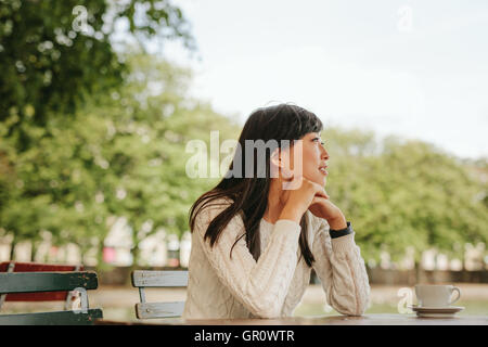 Giovane donna seduta al tavolo del bar con una tazza di caffè e guardando lontano. Femmina cinese in un momento di relax a outdoor cafe. Foto Stock