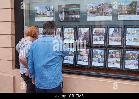 Coppia di età media cercando case in vendita, offerte immobiliari in finestra agente immobiliare a Londra, Inghilterra Regno Unito Regno Unito Foto Stock