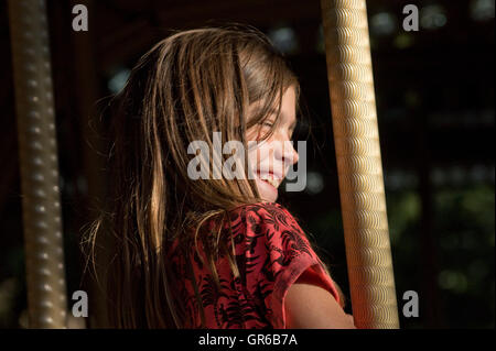 Ragazza giovane catturati nella luce solare e sorridente, seduto su un cavallo in una fiera Merry Go Round Foto Stock