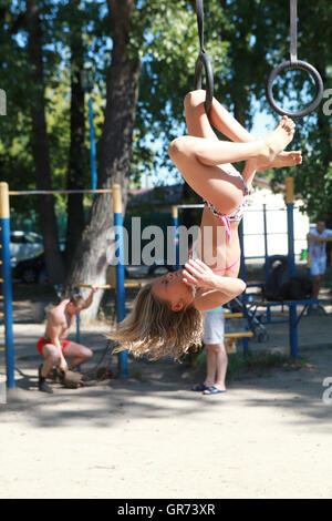 Una giovane ragazza gode il suo tempo su anelli di ginnastica, mentre suo padre è occupato il sollevamento pesi sul terreno dello sport a Kiev Hydropark area riverside Foto Stock