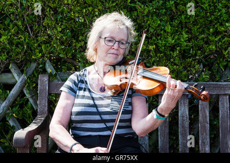 Donna matura con gli occhiali, seduti su una panchina nel parco suona il violino direttamente nella parte anteriore del visualizzatore. Siepe verde dietro di lei. Foto Stock