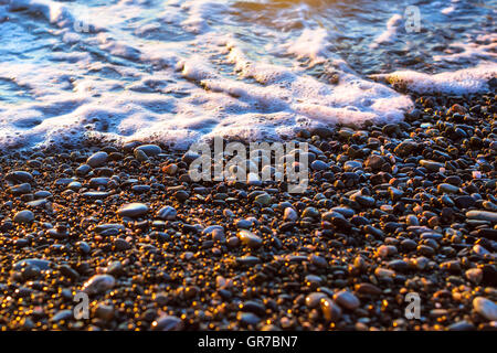 Schiuma di onde del mare si precipita a riva di ghiaia, riva della spiaggia è coperto di scintillanti pietre di sun. Sfondo di pietra, texture ciottoli Foto Stock