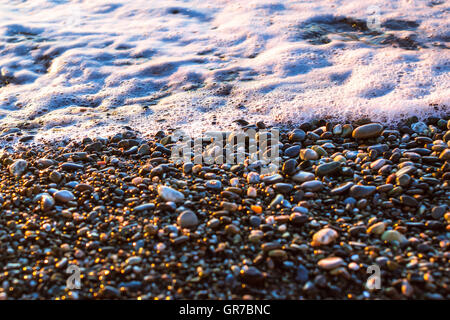 Schiuma di onde del mare si precipita a riva di ghiaia, riva della spiaggia è coperto di scintillanti pietre di sun. Sfondo di pietra, texture ciottoli Foto Stock
