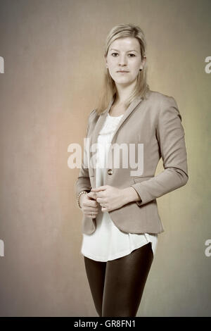 Giovane donna con capelli lunghi biondi che pongono in nero lucido e gambali Blazer prima di sfondo beige Foto Stock
