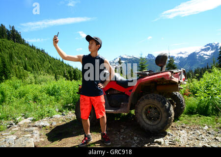 GenY maschio assume Selfie foto in natura Valle accanto a ATV utilizzando iPhone6 Onroute Smartphone attraverso il sentiero di ghiaia vicino a Whistler, BC Foto Stock