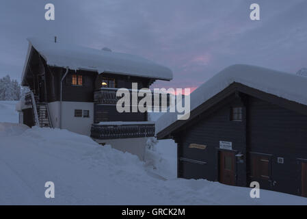 Dawn, Snowcovered percorso con due chalet a destra. Le due finestre sono illuminati. Sullo sfondo un grigio e leggermente rosa Sky.Pi Foto Stock