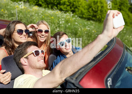 Amici guida in auto cabriolet e tenendo selfie Foto Stock