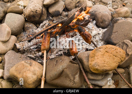 Salsicce per la cottura su un fuoco di campo su una riverside spiaggia ghiaiosa Foto Stock