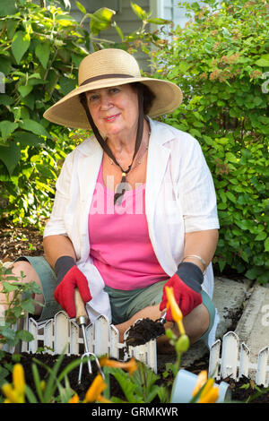 Senior donna sorridente in telecamera indossando cappello di paglia, abiti casual e guanti da giardinaggio mentre piantano fiori ornamentali in g Foto Stock