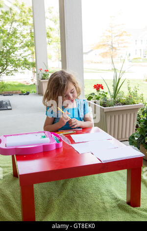 Carino bambina inginocchiati in un colorato rosso vivace colorazione tabella con matite su un outdoor patio coperto in s Foto Stock