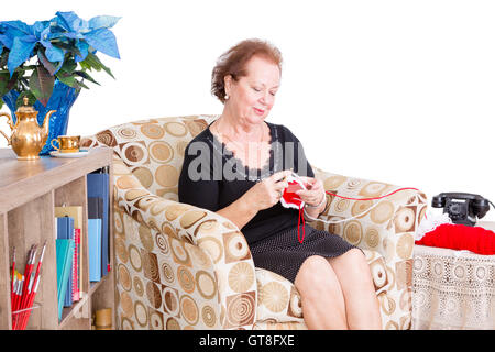 Nonna seduti a casa in una comoda poltrona nel soggiorno facendo la sua maglia con un look del piacere contenti Foto Stock