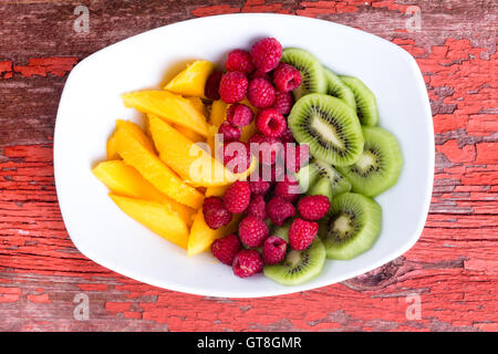 Mango e kiwi fette con lamponi in medio servita nella piastra bianca su tavola in legno rustico Foto Stock