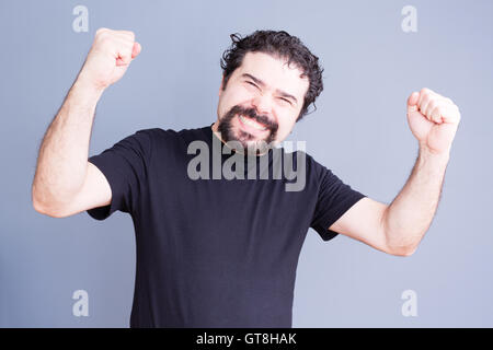 Bel singolo uomo barbuto in camicia nera tenendo i pugni fino con esultante espressione su sfondo grigio Foto Stock