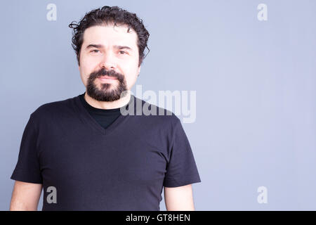 Vita Ritratto di uomo maturo con barba scura e capelli ricci vestita di nero T-Shirt e fissando con fotocamera Blank contenti Foto Stock