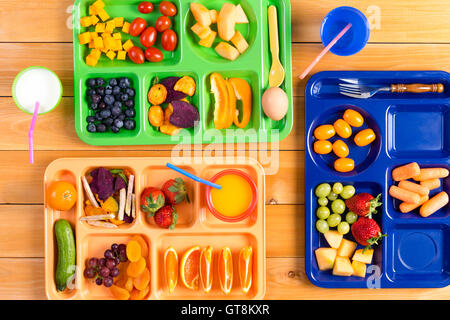 Vista dall'alto in basso di plastica colorato pranzo vassoi riempiti con fette e pezzi di frutta e verdura lungo il lato tazze di latte o j Foto Stock