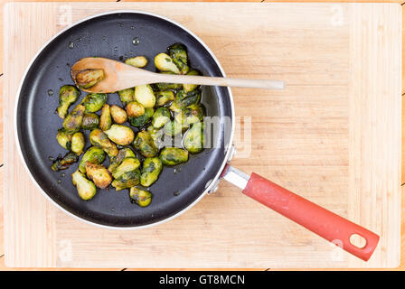 Top down prospettiva in prima persona di deliziosi i cavoletti di Bruxelles cotto in padella antiaderente con il cucchiaio di legno oltre il bordo di taglio Foto Stock