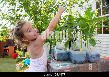 Carino bambina confrontando la sua altezza a fiori ella è piantare in plastica riutilizzate bottiglie di latte all'aperto vicino casa Foto Stock