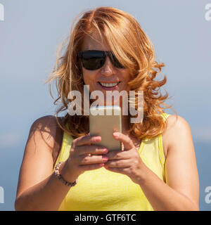 Donna con capelli biondi guardando lo schermo del suo telefono e sorridente. Ella riceve una buona notizia. Foto Stock