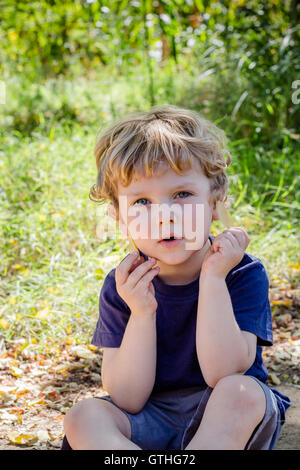 Piccola bionda ragazzo seduto in erba lunga guardando la telecamera trattenere foglie Foto Stock
