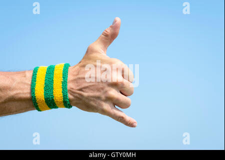 Atleta mano con giallo e verde i colori del Brasile con la fascetta da polso dando un classico shaka surfer segno contro il cielo blu Foto Stock