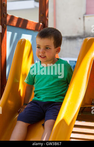 Adorabili poco ragazzo giocando su una diapositiva in un kids parco giochi esterno seduta al top con un caratteristico espressione come egli prepara t Foto Stock