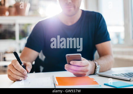 Imprenditore utilizzando il telefono cellulare mentre la scrittura nel diario. Immagine ravvicinata di man mano lavorando alla sua scrivania. Foto Stock