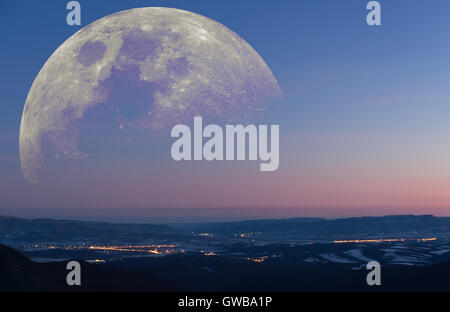 Fantastico paesaggio: inverno mattina presto vista dalle montagne di una valle con città di emettere luce con enormi derivanti luna Foto Stock