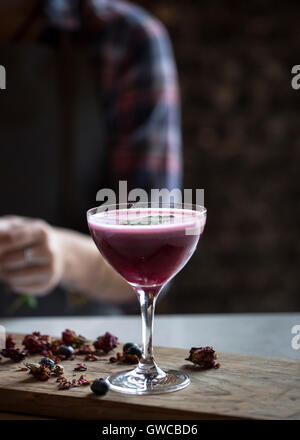 Una coupé di vetro riempita con mirtillo cocktail è fotografato in un bar con il barista nel retro al di fuori della messa a fuoco. Foto Stock