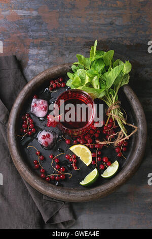 Ingredienti per rendere la limonata di cubetti di ghiaccio, ribes rosso bacche, calce, limone e menta, servita con bicchiere di succo di frutta sul vassoio di argilla wi Foto Stock