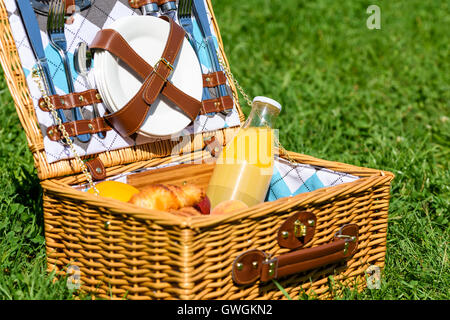 Cesto per pic-nic con succo di arancia bottiglia, mele, pesche, arance e croissant su erba verde in primavera Foto Stock