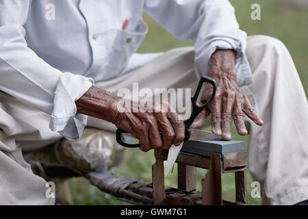 Affilare un paio di forbici con un macinacaffè a lama. Mani di un uomo asiatico anziano al lavoro. Thailandia Sud-est asiatico Foto Stock