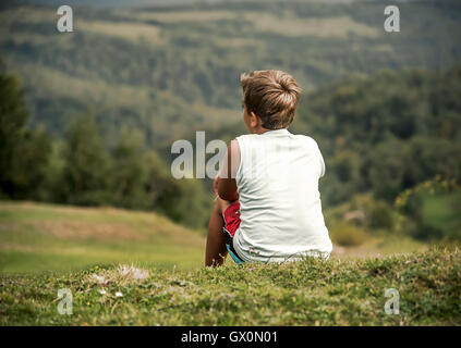 Ragazzo seduto su di una collina e si affaccia nella distanza. Infanzia, sogni, libertà - un concetto. Foto Stock